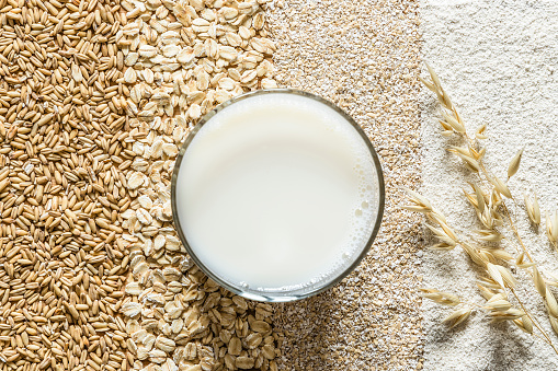 Vaso de leche de avena visto desde arriba y, de fondo, la avena en distintas fases: como cereal entero, procesado, salvado y en harina.