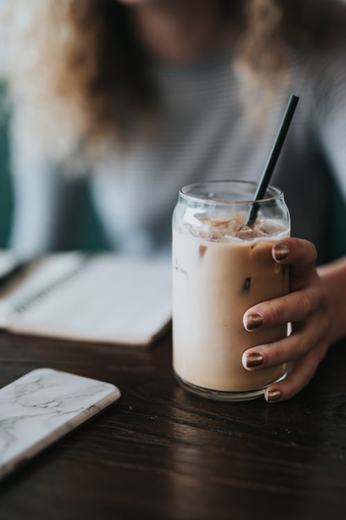 Mano de mujer sujeta un vaso con lo que parece café o té helado con leche de avena. Del vaso sobresale una pajita.