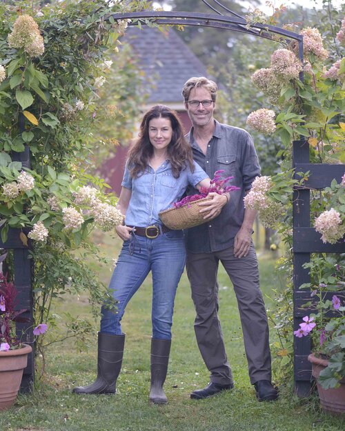 Pareja joven sonriente en la entrada de la granja, rodeados de flores y plantas. Ambos llevan vaqueros, camisa y botas de trabajo. Ella, en primer plano, sujeta con la mano izquierda una cesta con flores y se apoya sobre él.