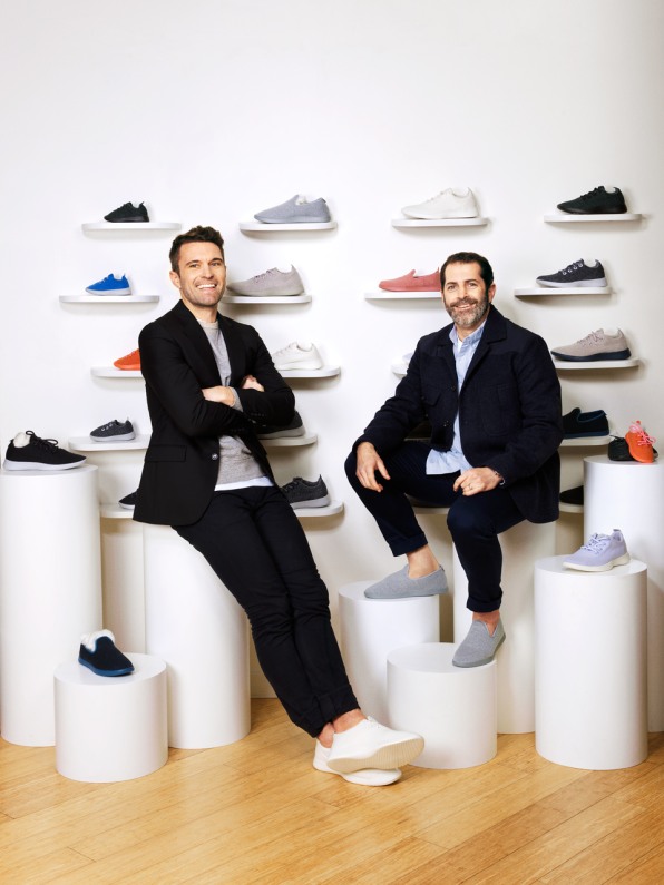 Dos hombres en traje y zapatillas sentados en una tienda de calzado con diversos modelos a su alrededor.